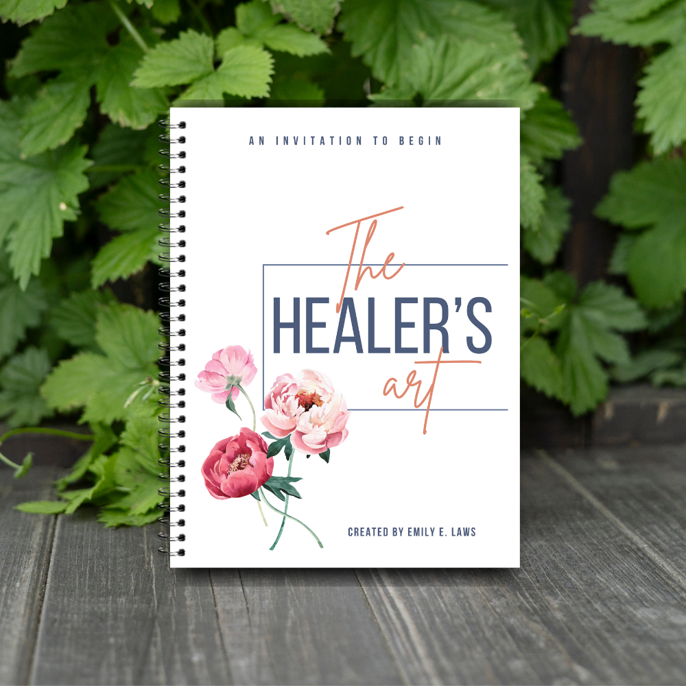 The Healer’s Art Workbook positioned on a rustic wood surface, surrounded by lush green foliage, evoking nature, grounding, and organic healing.