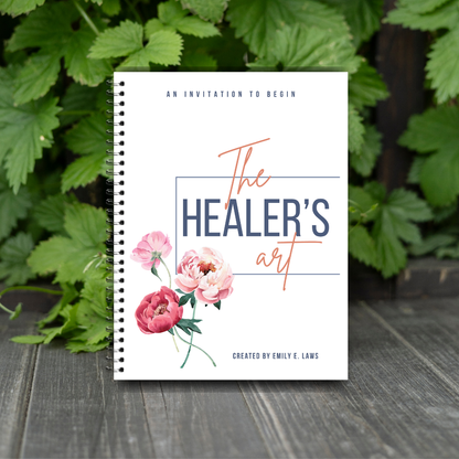 The Healer’s Art Workbook positioned on a rustic wood surface, surrounded by lush green foliage, evoking nature, grounding, and organic healing.
