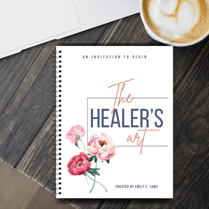 The Healer’s Art Workbook placed on a dark wooden desk beside a latte and a laptop, suggesting a cozy, mindful morning or creative workspace.